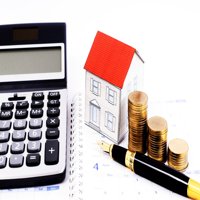 Real estate financial planning items A calculator, a small model house with a red roof, gold coins, and a fountain pen arranged on a calendar to represent home value and financial planning.